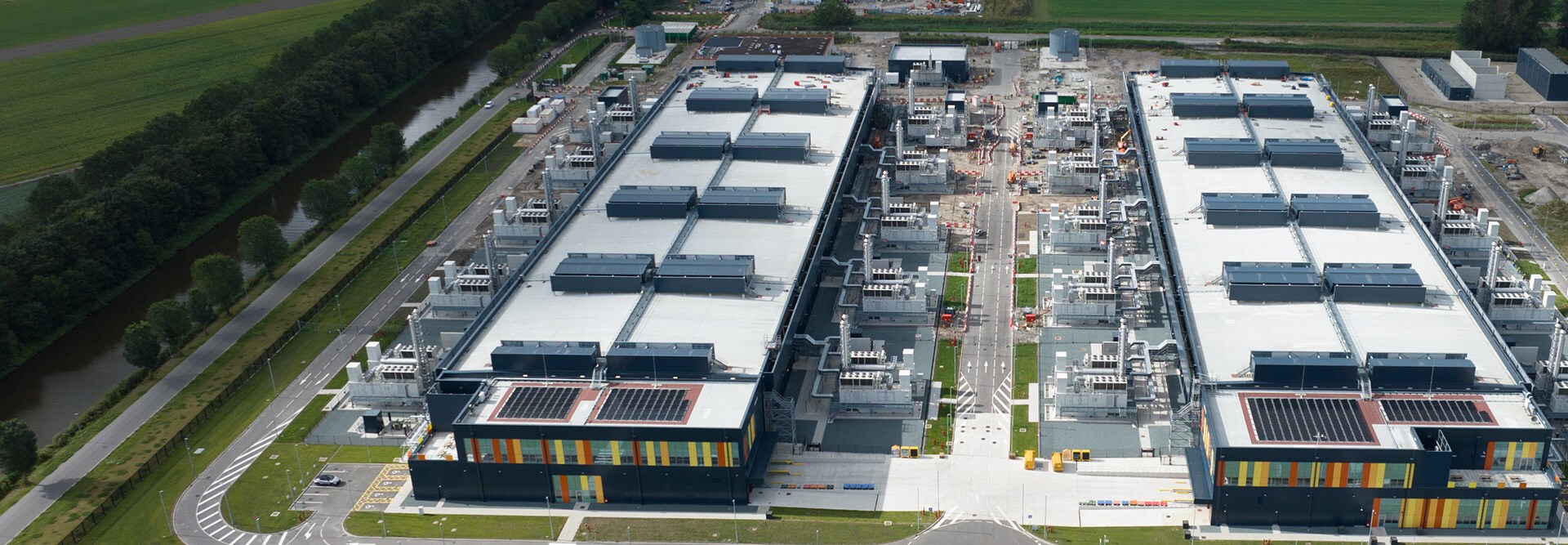 An aerial view of a large data center complex featuring two rectangular buildings with flat roofs covered in mechanical equipment and solar panels. 