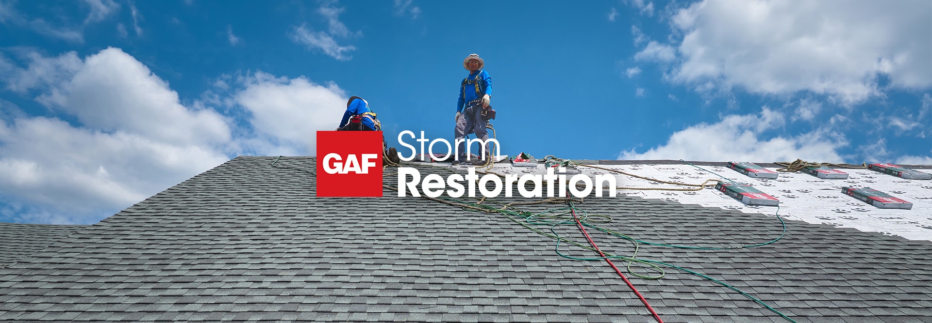 Two roofers working under a bright blue sky on a roof, with the GAF Storm Restoration logo overlaid on the foreground.