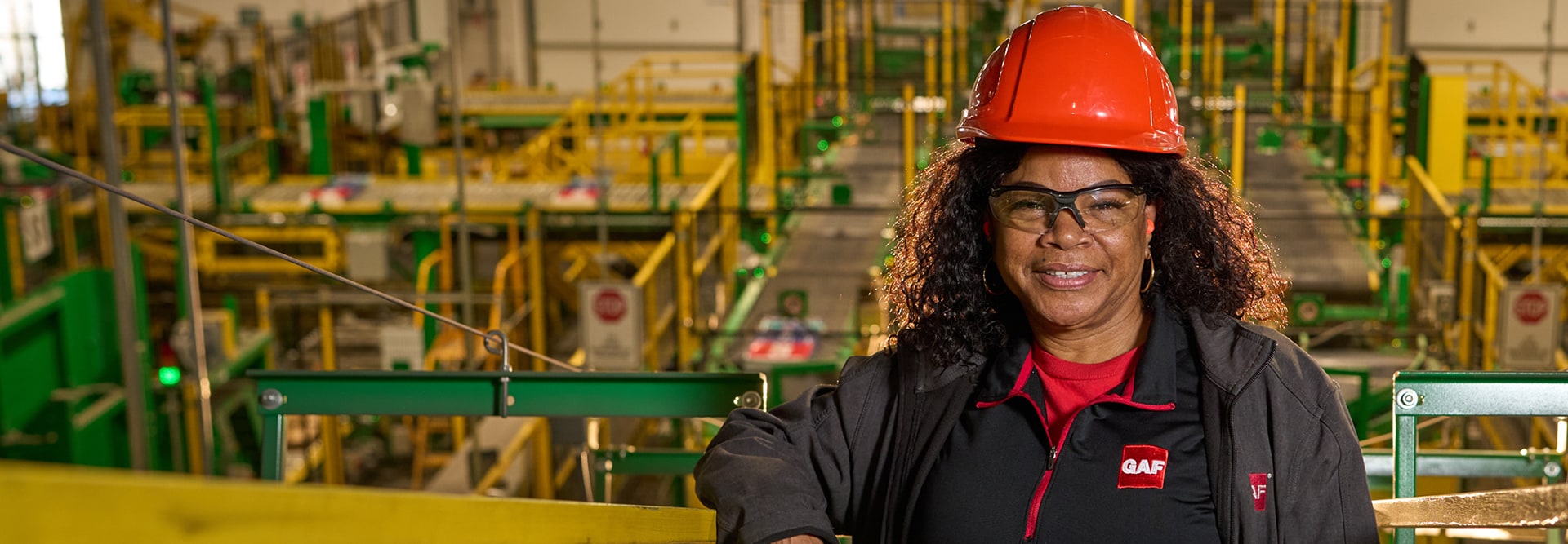 Female GAF manufacturing worker wearing an orange hard hat and safety glasses inside a factory facility.