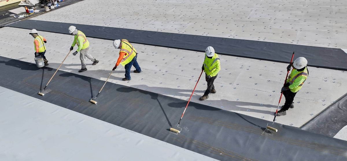Contractors rolling GAF bonding adhesive onto a flat roof
