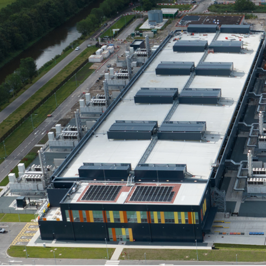 An aerial view of a large data center complex featuring two rectangular buildings with flat roofs covered in mechanical equipment and solar panels. 