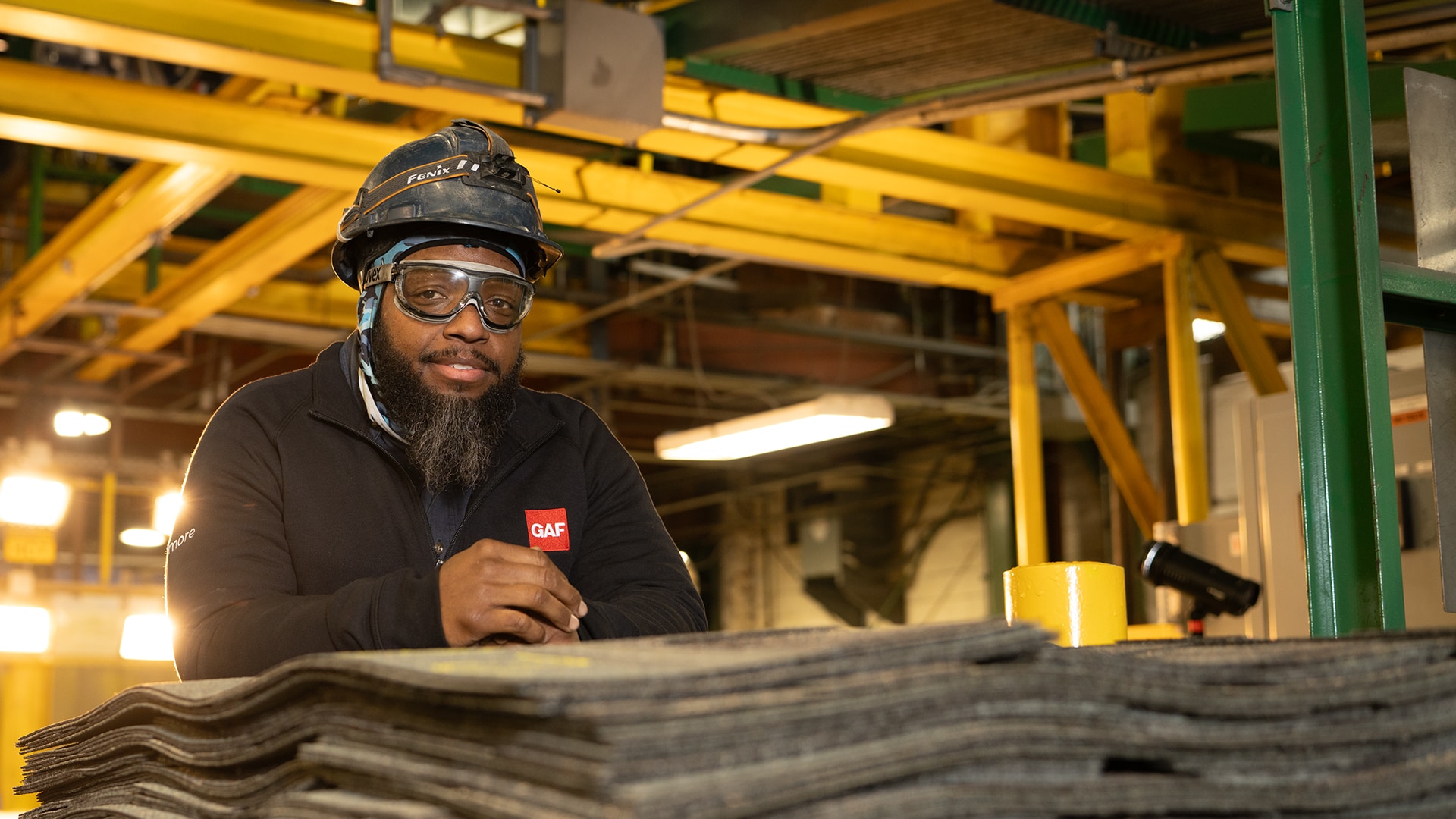 Male GAF factory worker wearing safety goggles and a hard hat standing beside stacked roofing shingles.