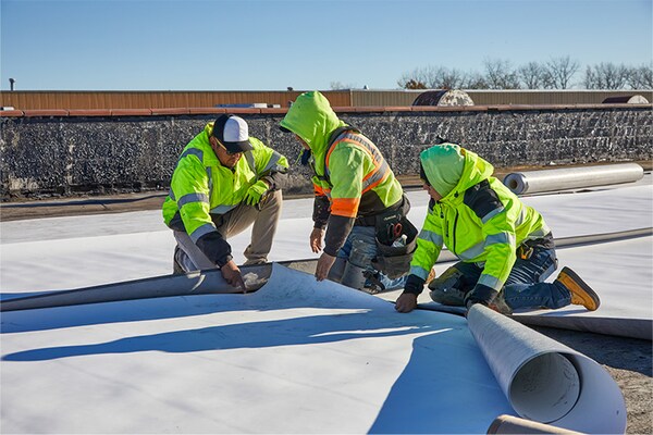 Three GAF certified commercial roofing contractors wearing high-visibility safety vests and jackets work together to unroll and align a large white thermoplastic roofing membrane on a flat rooftop.