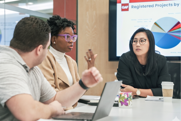 Three GAF professionals in an office setting reviewing commercial certifications data on a laptop and wall-mounted screen.