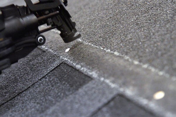 Close-up of a nail gun securing a GAF shingle to a roof, with a nail partially inserted along a line of GAF roofing shingles.