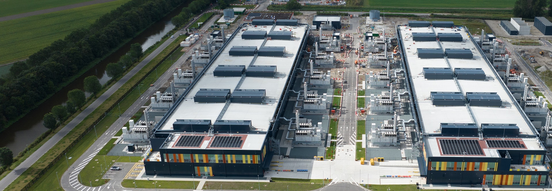 An aerial view of a large data center complex featuring two rectangular buildings with flat roofs covered in mechanical equipment and solar panels. 